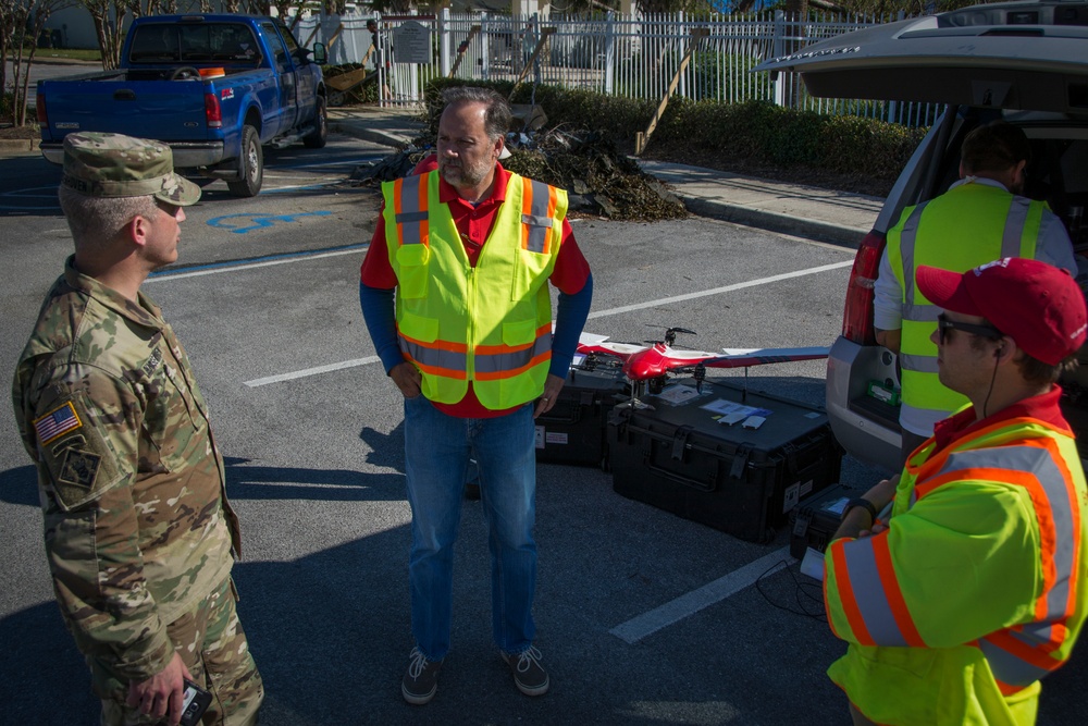 A U.S. Army soldier in OCP camouflage uniform is meeting with civilian contractors wearing high-visibility safety vests in a parking lot. The group appears to be discussing drone operations, as a red fixed-wing UAV is visible on equipment cases near the open trunk of a white SUV. The setting suggests a post-disaster or emergency response operation, supported by debris visible in the background.