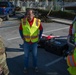A U.S. Army soldier in OCP camouflage uniform is meeting with civilian contractors wearing high-visibility safety vests in a parking lot. The group appears to be discussing drone operations, as a red fixed-wing UAV is visible on equipment cases near the open trunk of a white SUV. The setting suggests a post-disaster or emergency response operation, supported by debris visible in the background.