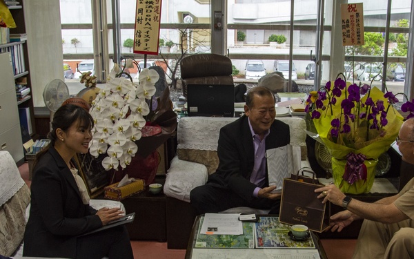 A formal meeting is taking place in what appears to be an official office, likely in Japan or Taiwan based on the visible Chinese/Japanese calligraphy and signage. Six individuals are seated around a glass-topped table displaying a map, engaged in what appears to be a diplomatic or official courtesy visit. A gift box is being exchanged between participants, and the room is decorated with floral arrangements including white orchids and purple flowers.