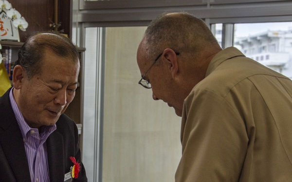 Two men are engaged in a formal card exchange, likely a Japanese business card (meishi) exchange ceremony. One man is wearing a black suit with a purple striped shirt and a lapel pin, while the other is dressed in a tan/khaki military-style uniform. The setting appears to be an official office or ceremonial room, with Japanese congratulatory signage visible in the background referencing the '18th generation Ginowan City' (第18代宜野湾市).