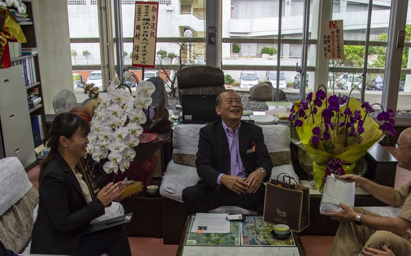 A formal meeting or reception is taking place in what appears to be an official office, likely in Taiwan or another East Asian location, with five individuals seated around a coffee table. A map is displayed on the table, and floral arrangements including white orchids and purple orchids are prominently visible. The atmosphere appears celebratory or diplomatic, with gift bags and tea cups present.