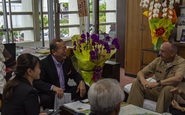 A formal meeting or courtesy call is taking place in what appears to be a Japanese official's office, with a U.S. military officer in tan uniform seated alongside civilians. The group of five people is gathered around a glass-topped table that has a map underneath it, engaged in what appears to be a friendly conversation. The setting includes decorative flower arrangements, Japanese signage, and office memorabilia consistent with a local government or civic office.