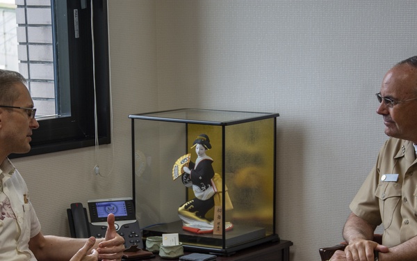 Two men are seated in an office setting engaged in conversation. One man is wearing a U.S. Navy uniform with visible ribbons and rank insignia, while the other is dressed in civilian attire with a floral shirt. The setting appears to be in Japan, evidenced by a traditional Japanese geisha doll displayed in a glass case between them.