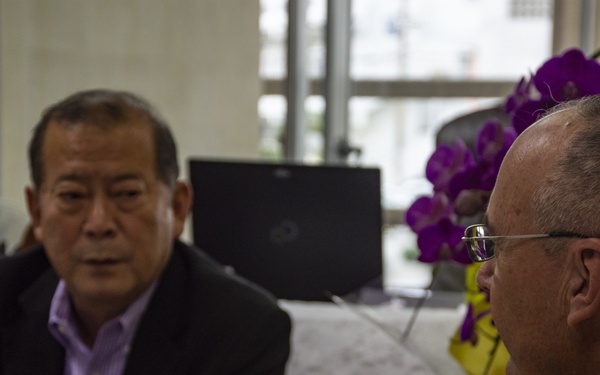 Two men are engaged in a face-to-face conversation in what appears to be an office or formal meeting room. One man, facing the camera, is wearing a black blazer over a purple shirt with a small red pin on his lapel. The other man, seen from behind, is wearing a tan/khaki shirt and glasses.