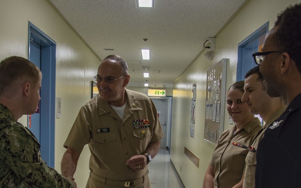 A group of U.S. Navy personnel are gathered in a hallway, with a senior officer in Service Khaki uniform shaking hands with a junior sailor in NWU (Navy Working Uniform) camouflage. Two additional sailors in Service Khaki uniforms stand nearby observing, along with a civilian in dark clothing. The setting appears to be a military facility, possibly a medical or administrative building based on the corridor design.