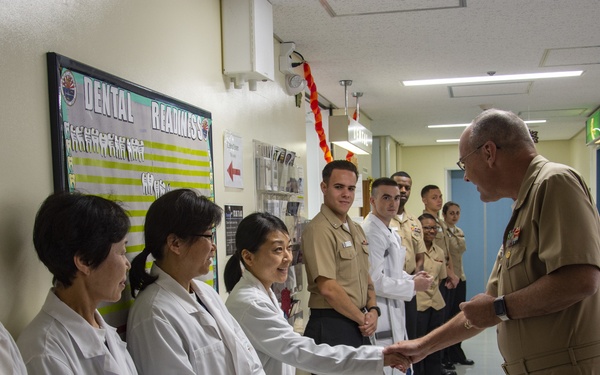 A senior military officer in tan/khaki uniform is greeting and shaking hands with staff in a dental facility corridor. The group includes personnel in white lab coats (likely civilian or medical staff) and uniformed military members. A bulletin board labeled 'DENTAL READINESS' with bilingual text (English and Japanese/Chinese characters) is visible on the wall, suggesting a U.S. military dental facility in Asia.