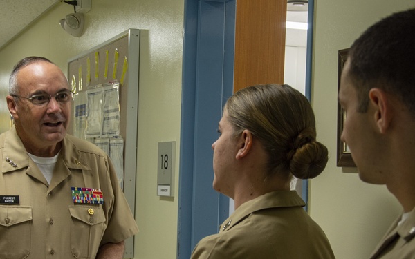 Three U.S. Navy personnel in tan/khaki service uniforms are engaged in a conversation in what appears to be a military medical facility hallway. The senior officer on the left, whose name tag reads 'FORREST FAISON,' displays multiple ribbons and what appear to be flag officer rank insignia, indicating a high-ranking admiral. He is speaking with two junior officers facing him.