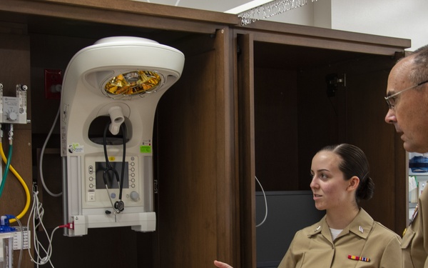 Two U.S. Navy personnel in khaki uniforms are standing in what appears to be a military medical facility, specifically near a neonatal or infant care station. The female officer is gesturing toward medical equipment while appearing to brief or explain something to the senior male officer. The setting includes a radiant warmer, infant bassinet, and various medical supplies and tubing mounted on a wooden cabinet unit.