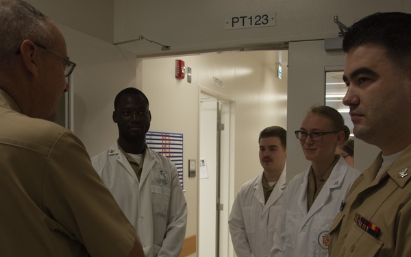 A group of military personnel are gathered in what appears to be a medical facility hallway. Two individuals wearing white lab coats stand alongside two others in tan military uniforms, engaged in conversation with a senior officer whose back is to the camera. The setting appears to be a military medical or hospital facility, indicated by the room designation 'PT123' visible above a doorway.