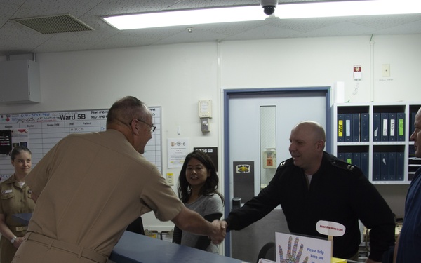 The image shows what appears to be a military medical facility, specifically a nursing station labeled 'Ward 5B.' A person in tan military uniform is shaking hands with a person in a dark uniform across a blue counter, while other personnel are present in the background. The setting appears to be a U.S. military hospital or medical center.