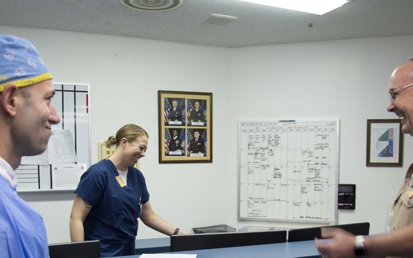 Three individuals are gathered at a medical facility nursing station, appearing to share a lighthearted moment. Two are in medical scrubs/surgical attire and one is in a U.S. military dress uniform with ribbons and insignia. The counter is decorated with autumn leaf cutouts and string lights, suggesting a seasonal holiday decoration.