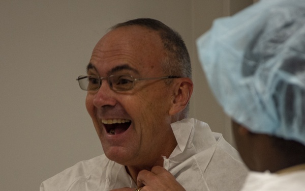 A middle-aged man wearing glasses and a white disposable protective gown is laughing broadly while another person in a blue hair net and white protective gown fastens or adjusts the collar of his gown. The interaction appears lighthearted and jovial, likely taking place in a medical, laboratory, or cleanroom environment. The man is wearing a gold ring on his right hand.