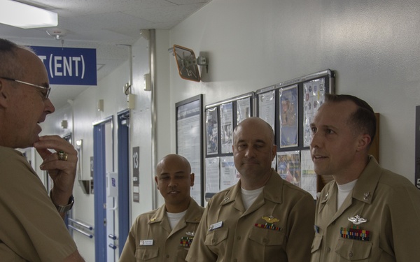 Four U.S. Navy personnel in tan/khaki service uniforms are engaged in conversation in what appears to be a military medical facility hallway. One senior officer faces three others, appearing to conduct a briefing or inspection tour. The setting shows numbered rooms and signage consistent with a naval hospital or medical center.