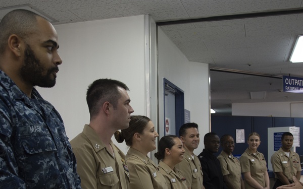 A senior U.S. Navy officer in khaki service uniform is addressing a group of Navy personnel in a medical facility hallway. The group includes sailors in both Navy Working Uniform (NWU blue digital camouflage) and khaki service uniforms. The setting appears to be a U.S. military outpatient medical facility, likely overseas based on visible bilingual signage.