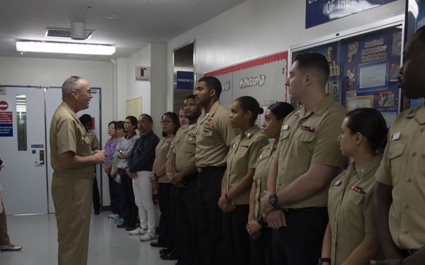 A senior military officer in U.S. Navy khaki uniform addresses a formation of sailors standing at attention in a hallway. The setting appears to be a military medical or administrative facility, with bulletin boards and signage visible on the walls. Civilian observers are visible in the background near a doorway.