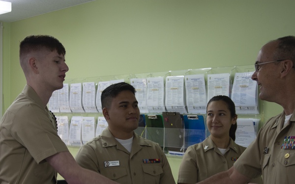 Four individuals in U.S. Navy tan/khaki uniforms are gathered in an office setting. A senior officer with multiple service ribbons is shaking hands with a younger, taller sailor, while two other sailors observe in the background. The interaction appears to be a formal greeting or recognition ceremony.