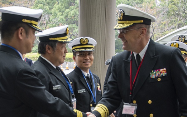 The image depicts a formal military greeting ceremony between naval officers from what appear to be two different nations. A senior Western naval officer wearing a US Navy-style uniform with extensive medal ribbons and a red lanyard is shaking hands with an Asian naval officer. Additional uniformed personnel are visible in the background observing the exchange.