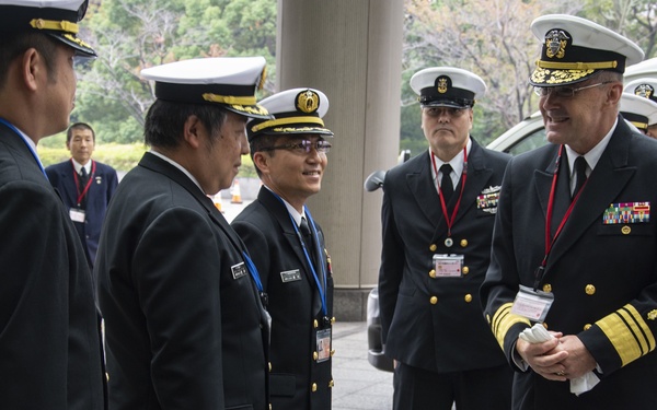 A group of naval officers in formal dress uniforms are gathered outdoors, appearing to engage in a diplomatic or ceremonial meeting. The group includes officers wearing what appear to be Japanese Maritime Self-Defense Force uniforms and United States Navy uniforms, suggesting a bilateral military exchange or official visit. The setting appears to be outside a formal government or military building.