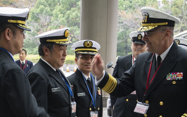 A group of naval officers in dress uniforms are engaged in conversation outside a building, appearing to be at an official meeting or diplomatic exchange. The group includes officers wearing what appear to be both U.S. Navy and Japan Maritime Self-Defense Force (JMSDF) uniforms, suggesting a bilateral military engagement. One senior officer on the right is gesturing while speaking to the others.