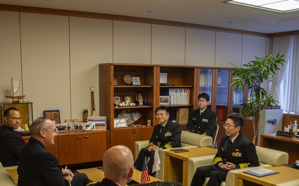 A formal military meeting is taking place in an office setting, with personnel in dark naval uniforms seated across from individuals in civilian and military attire. An American flag and what appears to be a Japanese flag are visible on a small table, suggesting a bilateral military or diplomatic meeting. The setting appears to be a senior officer's office, indicated by the formal furniture arrangement, display cases, and desk visible in the background.