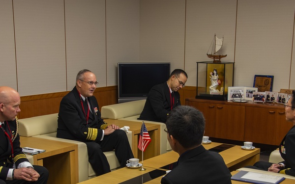 A formal military meeting is taking place in an office setting between U.S. Navy officers and what appear to be Japanese Maritime Self-Defense Force officers. The group is seated on cream-colored sofas arranged around low wooden coffee tables, with a small American flag visible on one of the tables. The setting suggests a bilateral military courtesy call or liaison meeting.