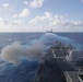 USS Spruance (DDG 111), foreground, fires a MK 45 5-inch gun while sailing in formation behind  USS John C. Stennis (CVN 74) during a live fire evolution.
