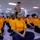 A large group of individuals wearing matching orange t-shirts and navy blue pants are seated on the floor in a large indoor facility. A uniformed military or correctional officer stands elevated at the back, overseeing the group. The setting and uniforms suggest a correctional boot camp, juvenile detention program, or military-style rehabilitation facility.