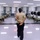 A military instructor in a tan uniform stands with hands clasped behind their back, overseeing a group of recruits in camouflage uniforms performing push-ups in two symmetrical rows. The setting appears to be a military barracks or training bay with bunk beds, black gear bags, and institutional fluorescent lighting. This appears to be a physical training or disciplinary exercise session.