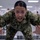 A person in U.S. Navy AOR2 woodland digital camouflage uniform is leaning aggressively toward the camera with mouth open and hands spread, appearing to shout or demonstrate an intense command presence. The setting appears to be a large indoor military facility, likely a training or processing center, with numerous other uniformed personnel visible in the background. The scene suggests a military training exercise or drill environment.