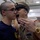 A military instructor or drill sergeant in a tan dress uniform with ribbons and decorations is yelling directly into the face of a young recruit wearing a navy blue t-shirt with a shaved head and glasses. The scene appears to take place inside a military training facility or gymnasium. This is a classic drill instructor confrontation typical of military basic training or JROTC/ROTC programs.