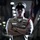 A military service member in dress uniform stands with arms crossed in a dramatic, low-lit corridor or hallway. The individual wears a white combination cover (cap) with a dark brim, a tan/khaki service uniform adorned with multiple ribbons, badges, and a red aiguillette. Flags are visible in the blurred background, suggesting an official military setting.
