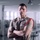 A male military service member in a tan/khaki uniform stands with arms crossed in what appears to be a classroom or office facility. He is wearing multiple service ribbons, a badge, a name tag, and a red aiguillette (decorative cord) on his right shoulder. His posture and expression convey a formal, confident demeanor.