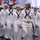 A large group of naval personnel dressed in white dress uniforms are marching in formation during what appears to be a military ceremony or graduation event. A female sailor in the foreground carries a ceremonial sword, indicating a leadership role. The event takes place indoors in a large hall decorated with flags and banners.