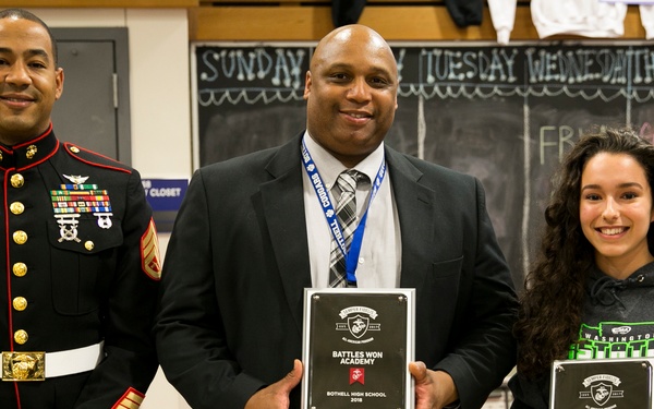 Three individuals pose for a photo in what appears to be a school gymnasium. A U.S. Marine in dress blues stands on the left, while a man in a suit holds a plaque reading 'Battles Won Academy - Bothell High School 2018,' and a young woman on the right holds a similar plaque reading 'Battles Won Academy - Liza Yusem 2018' along with a small box. The setting suggests an award ceremony recognizing the school and an individual student.