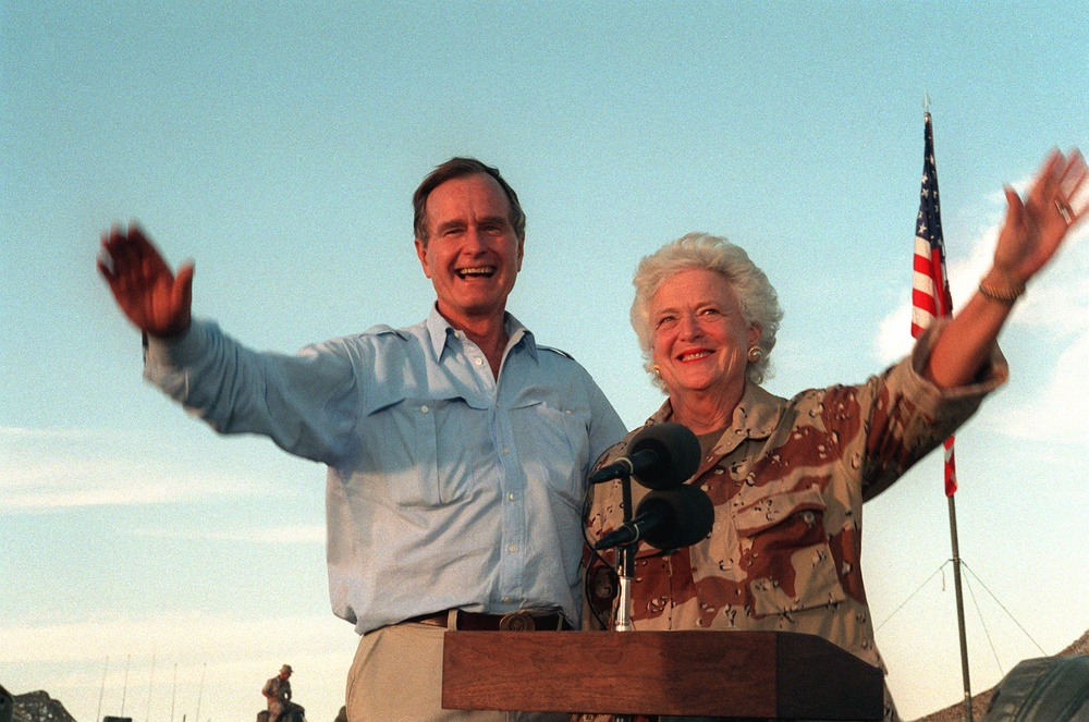 Two individuals, a man and a woman, stand together at a podium with microphones, both smiling and waving with arms outstretched. An American flag is visible in the background to the right. The setting appears to be an outdoor public or official event, likely during daytime with a clear sky.