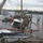 The image shows a post-storm marine salvage operation at a damaged marina or waterway. A crane barge with workers in safety gear is working to recover a partially sunken vessel, with an orange oil containment boom deployed around the wreck to prevent fuel spills. The surrounding area shows significant storm damage including downed trees, destroyed dock pilings, and multiple displaced or damaged boats.