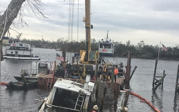 The image shows a post-storm marine salvage operation at a damaged marina or waterway. A crane barge with workers in safety gear is working to recover a partially sunken vessel, with an orange oil containment boom deployed around the wreck to prevent fuel spills. The surrounding area shows significant storm damage including downed trees, destroyed dock pilings, and multiple displaced or damaged boats.