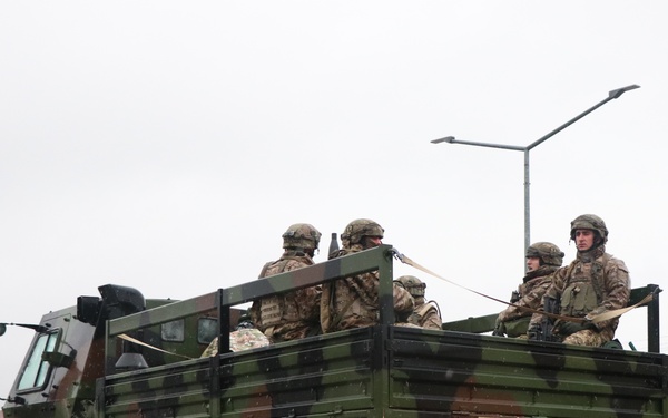 A military tactical truck with markings '7ATC' and 'PTF 311' is transporting several soldiers in full combat gear in its open cargo bed. The vehicle appears to be on a wet paved road within a military installation, suggesting rainy weather conditions. The soldiers are wearing camouflage uniforms and helmets, consistent with active duty or training operations.