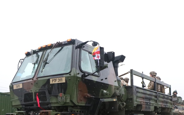 A military convoy is moving along a wet road in overcast conditions. The lead vehicle is a camouflage-painted heavy tactical truck marked 'PTF 311' and '7ATC', carrying armed soldiers in the flatbed. A small flag consistent with the Georgian national flag is visible mounted on the lead vehicle.