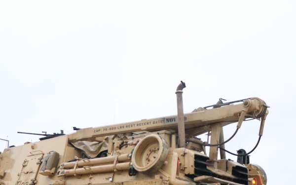 The image shows a desert-tan painted M88 Recovery Vehicle (ARV), a tracked armored recovery vehicle used by the U.S. Army, positioned next to what appears to be a flatbed transport trailer. The vehicle displays visible markings including 'L 11' on its side and a crane/boom arm extended over the rear. The setting appears to be a military installation or staging area under overcast conditions.