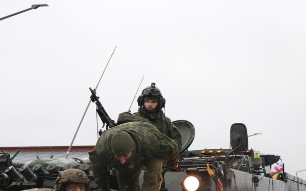 The image shows military personnel operating near an armored personnel carrier (APC), likely an M113 variant, in a wet outdoor environment. At least three soldiers are visible, wearing different camouflage patterns suggesting a multinational or joint exercise. The scene appears to take place on a military installation or base, with vehicles staged on a wet road.