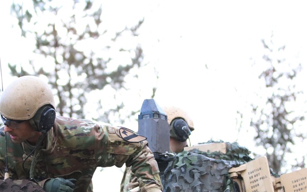 Two military personnel in camouflage uniforms and tan combat helmets are working on top of an armored military vehicle. They appear to be adjusting or deploying camouflage netting over the vehicle's exterior. The vehicle is tan/desert-colored and features warning labels and communication equipment.