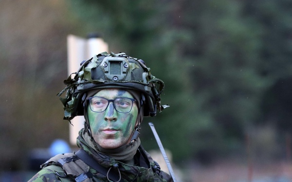 A fully equipped soldier wearing green camouflage face paint and a tactical helmet stands in an outdoor environment. The soldier is wearing a Lithuanian flag patch on the left sleeve, indicating affiliation with the Lithuanian Armed Forces. The image appears to be taken during a military exercise or patrol operation.