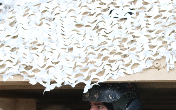 A soldier is partially visible inside or beneath a military vehicle, wearing a combat helmet with communications headset. The vehicle is covered with a winter/desert camouflage netting in white and tan patterns. The soldier appears to be in an observation or operational position within the vehicle hatch.