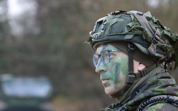 A soldier in full combat gear is shown in profile view, holding a military radio/handheld transceiver with an extended antenna. The individual is wearing camouflage face paint in green tones, a tactical helmet with netting, and a camouflage uniform with a visible shoulder patch. The setting appears to be an outdoor military exercise or operational environment.