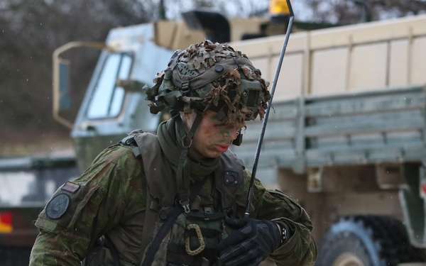 A soldier in full combat gear is operating a radio handset in what appears to be a field training exercise or operational environment. The soldier is wearing camouflage uniform with a camouflage-netted helmet, tactical vest, and face paint, suggesting a field operation. Military vehicles, including what appear to be tactical trucks, are visible in the background.