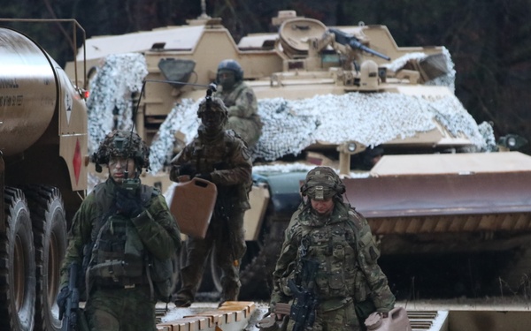 The image shows multiple soldiers in different camouflage uniforms operating in a military field environment alongside armored vehicles. At least two distinct uniform types are visible, suggesting a multinational or joint military exercise. Soldiers are carrying jerry cans and weapons while moving around a flatbed trailer area near military vehicles.
