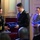 Maryland Guard, Bosnia-Herzegovina armed forces celebrate 15-year partnership A military ceremony is taking place in a formal indoor venue where a female U.S. Army officer is presenting a Certificate of Appreciation to a male officer in a dark naval-style uniform. An American flag is visible in the background, and a third uniformed individual stands nearby holding a blue folder, likely reading from a program. An audience of uniformed personnel is seated in the foreground.