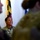 Maryland Guard, Bosnia-Herzegovina armed forces celebrate 15-year partnership A female military officer in camouflage uniform is smiling broadly, appearing to be engaged in a conversation or group interaction. She is surrounded by other uniformed personnel, with the Maryland state flag visible in the background. The setting appears to be an indoor military facility or office.