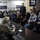 Maryland Guard, Bosnia-Herzegovina armed forces celebrate 15-year partnership Two U.S. Army soldiers in camouflage uniforms are seated at a table facing a group of young civilians in what appears to be a community meeting or outreach event. A young man in a dark suit stands at the center, likely serving as a moderator or translator. The setting appears to be a cultural or community hall with framed photographs on the walls.